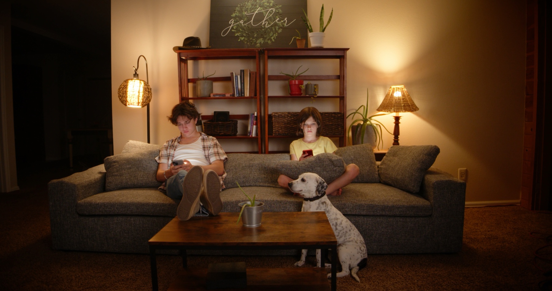 Two siblings sit on a couch with their dog, both on their phones.