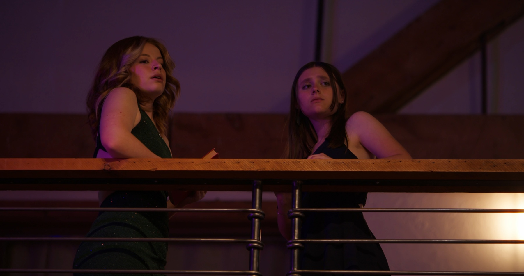 Two girls lean on a railing at a dance, lit in purple and gold.
