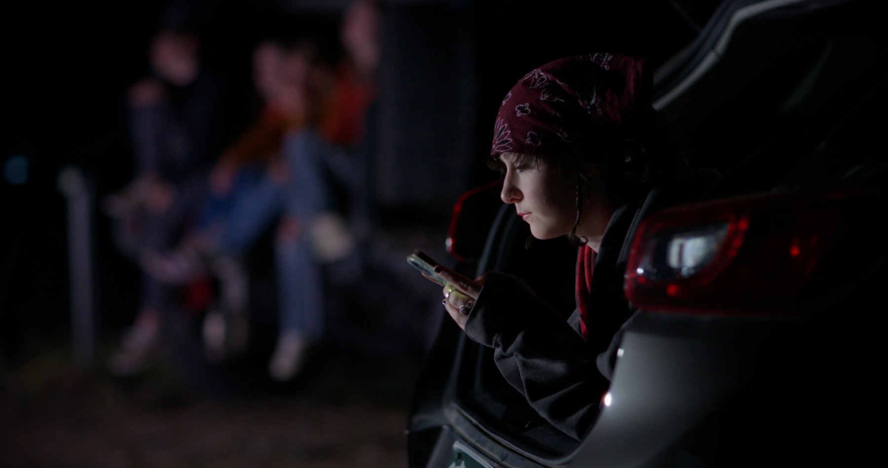 A girl in a bandana sits in a car trunk at night, lit by her phone.