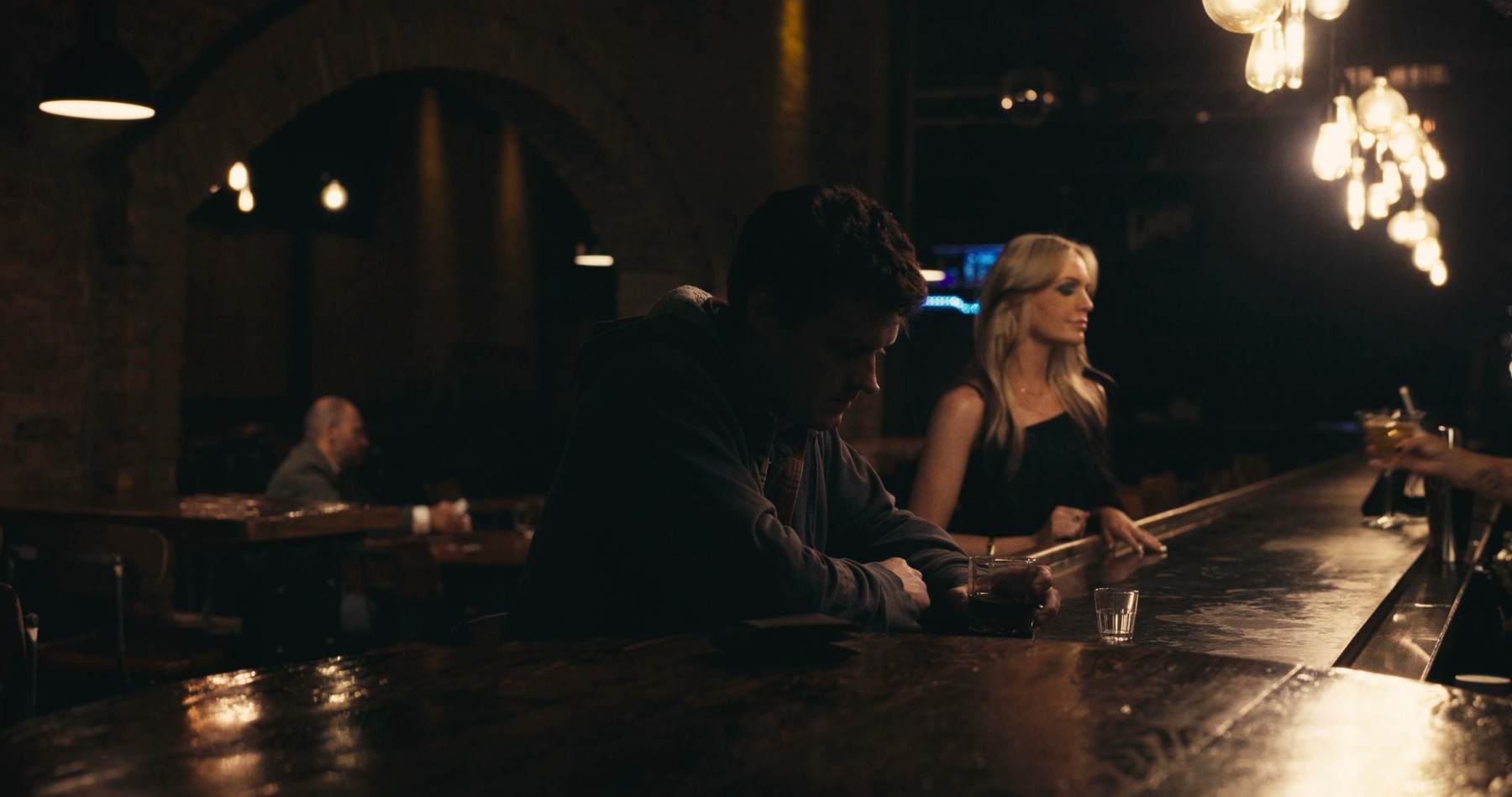 Milo and a woman sit across a table under warm bar lights.