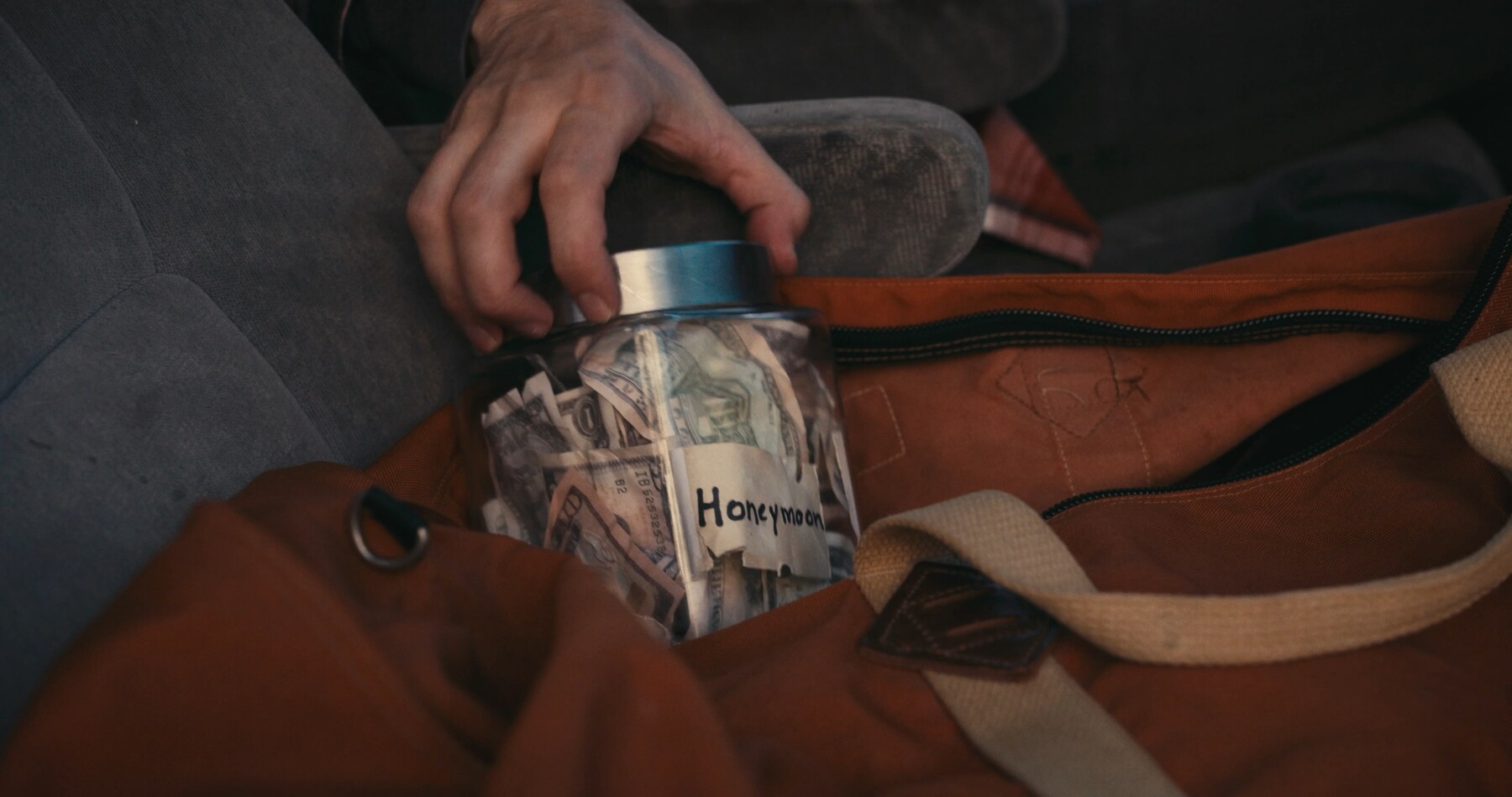 A hand tucks cash into a jar labeled Honeymoon.